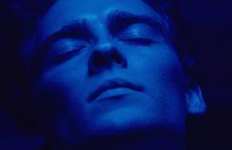 A close-up of a young man with his eyes closed in pleasure, he is cast in a deep blue light.