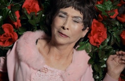 An elderly woman wearing a pink fluffy shawl lies in a bed of fake roses.