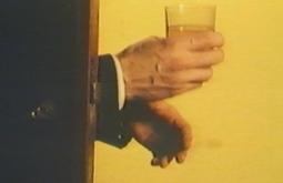 A pair of hands appear behind a door, one had holds a glass of water.