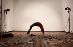 In a white walled gallery a person does a bridge pose, there aer two spotlights on either side of them. Paper is scattered on the floor.