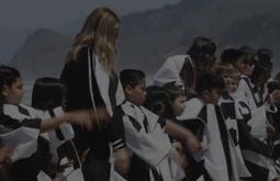 A group of women and children stand on the coast wearing full body length black & white robes swinging their arms.