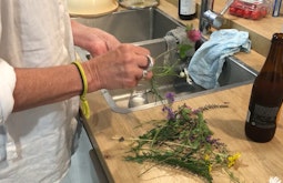 Standing by a sink someone trims the stems of freshly picked flowers. An open bottle of beer stands beside the sink.