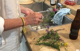 Standing by a sink someone trims the stems of freshly picked flowers. An open bottle of beer stands beside the sink.