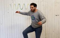 A man stands in front of a dirty white wall of a barn dancing and stomping.