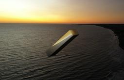 A digitally rendered metal oblong flies above the sea while the sun sets.