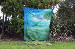 A large piece of fabric painted with an image of islands and trees hangs from a cord suspended amongst native New Zealand bush.