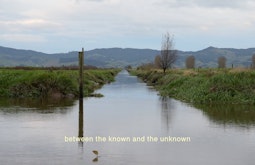 A calm river flows amongst grassy farmland. There are moutnains in the background and a fish jumping in the foreground. Subtitles read "between the known and unknown"