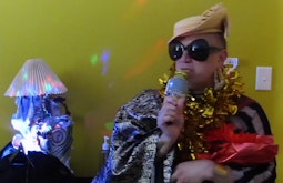 A person is singing into a gold microphone while wearing gold tinsel. There is a colourful party light shining beside them.