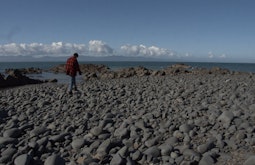 A man in a red and black flannel and beanie is walking along rocks at the coast. There are mountains and clouds in the distance.