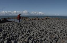 A man in a red and black flannel and beanie is walking along rocks at the coast. There are mountains and clouds in the distance.