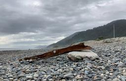 A rusty and warped iron beam rests on coastal rocks. A digger is working in the background.