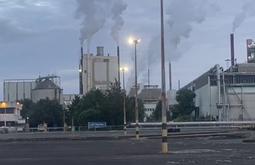 A large factory with smoke stacks billows gasses into the air. Flood lights are in the foreground shining over bare concrete.