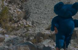 A person wears a large blue furry teddy bear costume as they walk amongst dry rocks.