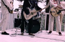 A four person band plays on a white wood deck. Three people are playing guitars and one is playing the drums.