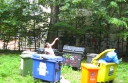 A person stands in a small park encircled by wheelie bins of various colours.