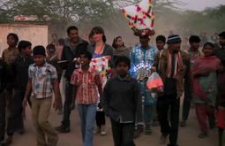 In india a crowd of people surround a performer dressed in iridescent blue with a large headpiece.