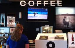 At a cafe till a person stands waiting to order. A big sign reading coffee hangs from the ceiling.