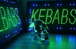 A chrome skull sits on a table between two neon signs that read kebabs.
