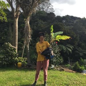 Ahilapalapa stands outside, holding a large plant with plastic over it’s roots, smiling. They are wearing a yellow t-shirt which reads “PASIFIKA”, red pants and gumboots.