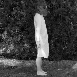 A black and white still shows Aliyah Winter standing outside on grass. Her arms are held out by her sides, extended slightly as if waiting for something or performing an action. The image is taken from a side profile perspective. She wears a long white dress shirt, standing on slightly wet grass in front of a hedge.