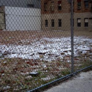 An area of rubble, behind a metal fence, covered in a white substance which could be snow. A brick building is in the background.