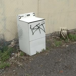 A discarded and graffitied washing machine sits near pieces of rubbish in a vacant car park or lot near a wire mesh fence.