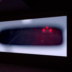 A film is projected onto a wall, in it we see the brake lights of a small car in a blurry rear-view mirror.