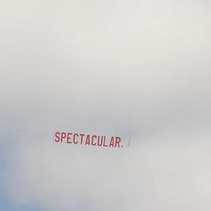 A large red ariel banner is flown through the air, towed from an out of the shot aeroplane. The text on the banner reads "SPECTACULAR." in large red lettering against a cloudy blue sky