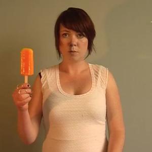 Erica holds up a bright orange popsicle in front of a plain wall. They wear a white singlet and a deadpan facial expression