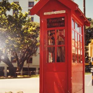 A scanned image of a old photo of a person standing in a red phone box, holding a phone to their head.