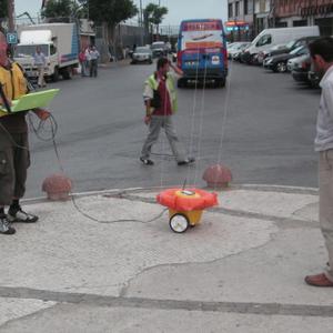 A street scene with people gathered round a small yellow object with wheels that is attached to four thin strings.