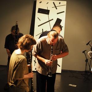 A group of people play woodwind instruments in a gallery. At centre rear, a large rectangular graphic score leans against the wall. On the floor are are two much smaller rectangles. All of the performers look down as they play, as if referencing the rectangles on the floor, which act as graphic scores.