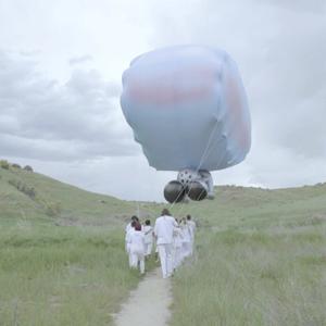 A group of people in white outfits carry the strings of a giant balloon through a grassy landscape