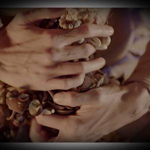 A close up of two hands clutching a string of beads and shells. Linear tattoos are visible on the figure's fingers and wrists