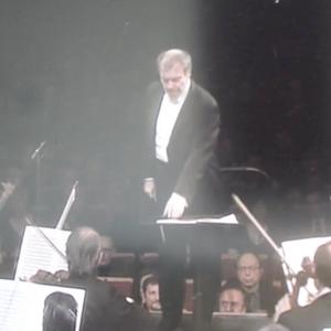 A conductor stands in front of an orchestra wearing a suit