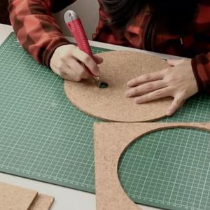 A person sits at a desk and uses a craft knife to carve a circle out of a cork board.