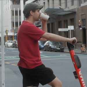 Max is on a segway, waiting to cross the street, drinking out of a white takeaway cup.