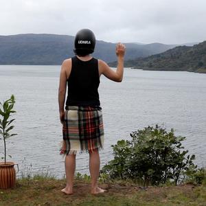 A person stands on the shoreline above a harbour. At their left is a potted plant, beside that is a naturally planted scrub bush. The person wear a tartan blanket, a black singlet and a black motorcycle crash helmet with the capital letters UDNRA printed on the back. Their right arm is raised as if in greeting or solidarity