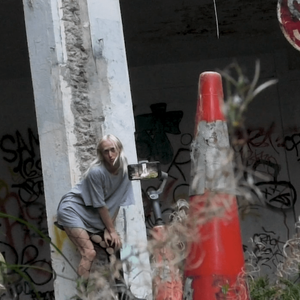 The artist leans against a tall concrete poll whilst a phone on a tripod is filming them. They are in an abandoned car park with wet concrete and weeds.