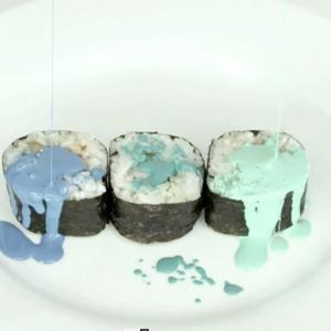 Three sushi rolls on a plate are drizzled with green and blue paint