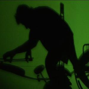 The silhouette of a figure standing against a green background. A chair is also silhouetted and other instruments.