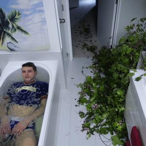 A still from Alex Plumb’s work shows a person in the bath, fully clothed with a blank expression on their face. The image is taken from a high angle, looking down into the entire bathroom and down the hallway of the house. A large overgrown plant spills messily through the bathroom and down the corridor. The image feels airy, suspenseful, uncanny.