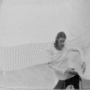 An image of a dancer near a wall mounted radiator. The image is shot onblack and white 8mm film, is grainy and appears to be warped by superimposition of two stills from the same film