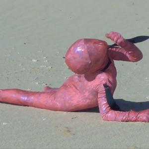 A figure dressed in a full-body holographic pink morph suit crawls along the sand on a sunny beach. They are potentially being walked like a dog or pulled along. This is shown by a black piece of fabric around their neck leading outside of the image to something or someone we can not see.