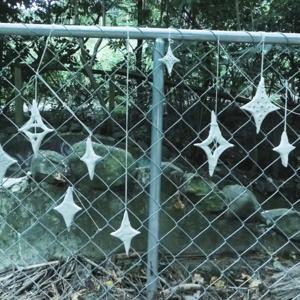 A series of 9 Matariki stars hang from by stringa chainlink fence. The stars are hand made and some have holes.