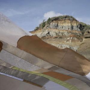 Flowing fabric flows gently with the wind in an outdoor location with a rock cliff behind it. The fabric piece is a patchwork made up of different beige coloured material, all with slightly different textures and transparency.