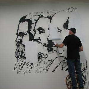 A person stands on a chair to paint over portraits of three figures painted in black paint.