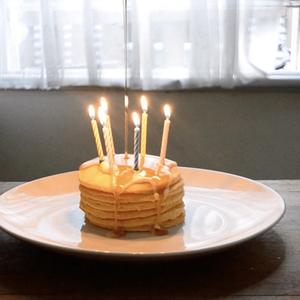 A stack of small pancakes decorated with birthday candles is dripping with honey on na plate. The lit pancakes sit alone on a table in a flat, behind the table is a window with netted curtains.