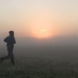 A person is jogging across a grassy field in the early morning. The landscape is covered in thick fog, blurring the horizon line. We can see the red glow from sunrise. The person running appears slightly mid-air, in between steps. The dawn makes them look like a silhouette on the land. The grass is covered in dew or frost.