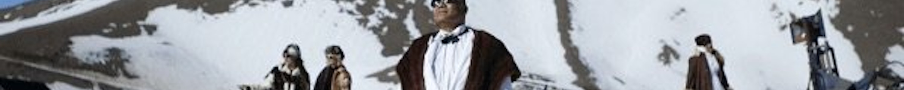 In this still of Christopher Ulutupu's video work the artist's father stands on a sunny, snowy mountain peak wearing a dapper white suit, sunglasses and fur throw, looking up towards the sun shining. The artist’s mother and sisters are in the background all wearing glamorous clothing in the bright snow
